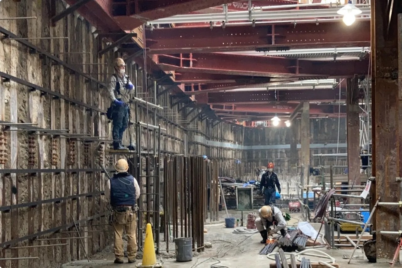 地下工事現場で、ヘルメットとマスクを着用した作業員数名が、鉄骨と土留め壁の前で作業に従事している様子。